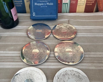 May include: Six silver-colored coasters with an intricate design. The coasters are arranged in a semi-circle on a striped surface. A blue box with the text "Mappin & Webb" is in the background.