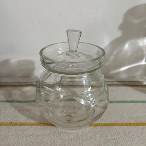 May include: A clear glass jar with a lid and a small handle. The jar has a round body with etched floral designs. The lid is also clear and fits snugly on top. The jar is sitting on a striped surface.