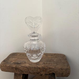May include: A clear glass perfume bottle with a heart-shaped stopper. The bottle has a round body with etched floral designs. It sits on a rustic wooden stool. The stopper is clear and has a leaf-like pattern.