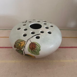 Puede incluir: Un pequeño jarrón de cerámica blanco con forma redonda y un anillo de agujeros alrededor de la parte superior. El jarrón está decorado con un diseño floral verde, marrón y amarillo.