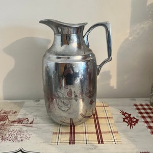 Könnte beinhalten: Eine silberne Metallkanne mit rundem Körper und Henkel. Die Kanne steht auf einem karierten Untersetzer.