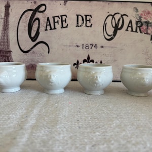 Op de afbeelding: Vier kleine, witte keramische kommen met een leeuwenkopontwerp. De kommen zijn in een rij gerangschikt tegen een achtergrond met de Eiffeltoren en de woorden "Cafe de Paris 1874."