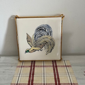 May include: A square ceramic tile with a rooster illustration in flight, in cream, yellow, blue, and brown. The tile is set in a gold-coloured metal frame, creating a decorative accent. The rooster design is detailed and artistic.