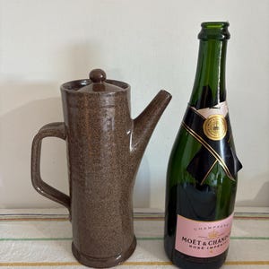 Puede incluir: Una jarra de cerámica marrón con asa y boquilla, junto a una botella de champán verde con una etiqueta rosa. La jarra tiene una textura moteada. La botella de champán tiene una etiqueta negra y dorada.