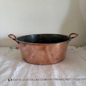 Könnte beinhalten: Eine Kupferschüssel mit zwei kleinen Griffen. Die Schüssel hat eine abgerundete Form und ein dunkles Inneres. Die Außenseite hat eine glänzende, rötlich-braune Farbe. Die Schüssel steht auf einer weißen Tischdecke mit Blumenmuster.