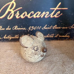 Könnte beinhalten: Eine gesprenkelte Keramik-Mausfigur in Beige mit großen Ohren und einer braunen Nase. Die Maus hat einfache Gesichtszüge und steht auf einer Jutefläche. Der Hintergrund zeigt ein Vintage-Schild mit der Aufschrift "Brocante".