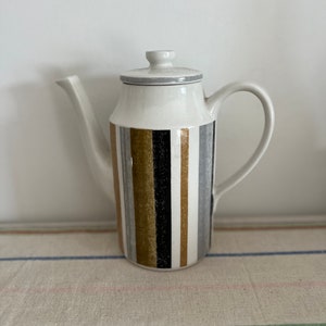 May include: A white ceramic coffee pot with a striped design in black, gold, and gray. The pot has a lid and a handle.