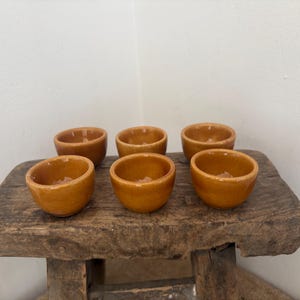 May include: A set of six small, round, ochre-colored ceramic bowls with a smooth, glossy finish. Each bowl features a simple, open design with a consistent depth and width, arranged in two rows of three on a rustic, weathered wooden surface.