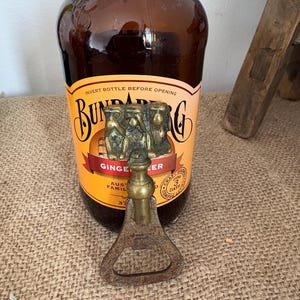 May include: A vintage bottle opener with a bronze handle and a decorative top with three figures. It is placed in front of a brown glass bottle with a "Bundaberg Ginger Beer" label. The bottle opener is made of metal.