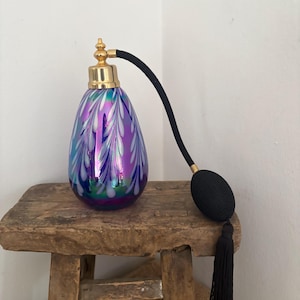 Può includere: Un flacone di profumo vintage con un corpo in vetro iridescente a forma di pera nei toni del blu, viola e verde. Presenta un colletto in metallo color oro e un atomizzatore nero con un nappa abbinata. Il flacone è esposto su uno sgabello di legno rustico.