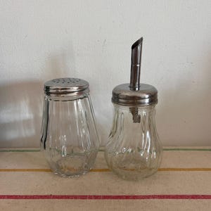 May include: Two clear glass sugar dispensers with silver lids and a silver spout on one of the dispensers. The dispensers have a ribbed design and are sitting on a striped tablecloth.