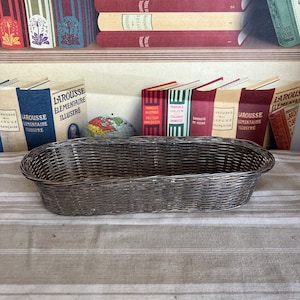 Könnte beinhalten: Ein silberfarbener, ovaler Drahtkorb auf einer gestreiften Leinentischdecke. Der Korb ist leer und bereit, Gegenstände aufzunehmen.