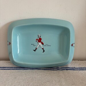 May include: A light blue ceramic tray with rounded corners. The tray features a central illustration of a man in a red coat and white hat, with the text "BORN 1820 - STILL GOING STRONG". Decorative elements are on the sides.