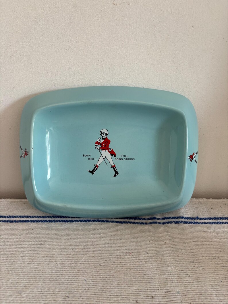 May include: A light blue ceramic tray with rounded corners. The tray features a central illustration of a man in a red coat and white hat, with the text "BORN 1820 - STILL GOING STRONG". Decorative elements are on the sides.