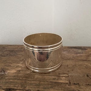May include: A shiny, silver napkin ring with a cylindrical shape. The ring has two thin, horizontal lines etched around the middle. The interior of the ring is a warm, golden color. The napkin ring sits on a weathered, wooden surface.