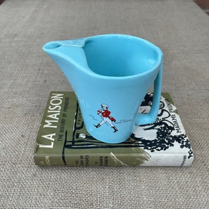 May include: A light blue ceramic jug with a handle and a spout. The jug features a graphic of a man in a red coat walking with a cane and the text "Still Going Strong".