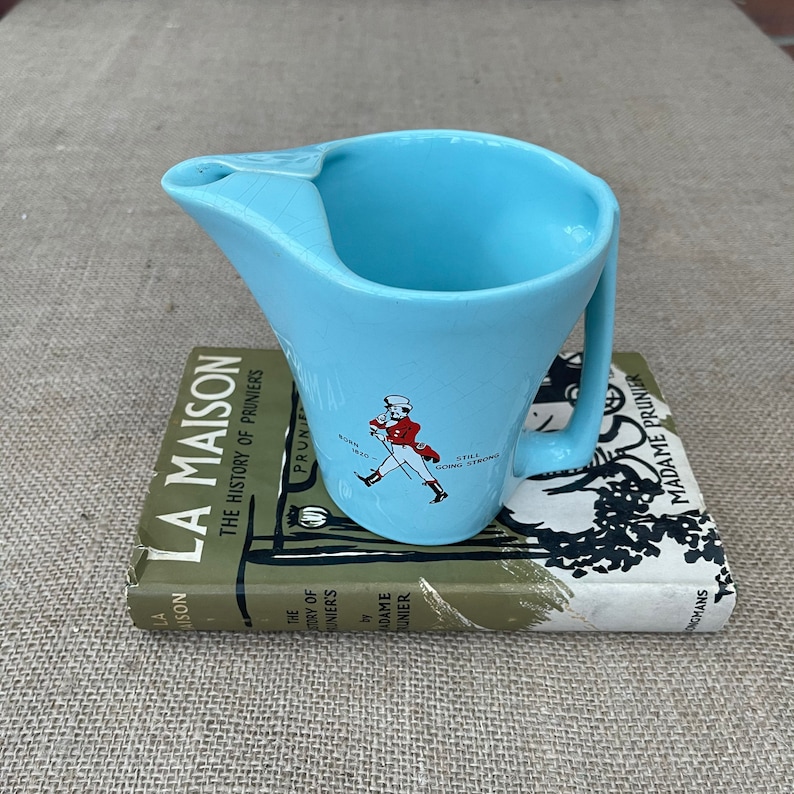 May include: A light blue ceramic jug with a handle and a spout. The jug features a graphic of a man in a red coat walking with a cane and the text "Still Going Strong".