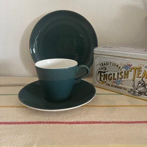 Puede incluir: Un juego de taza y platillo de té verde azulado con borde blanco. La taza de té tiene un interior blanco. La taza y el platillo están sobre un mantel a rayas. Una lata de "Traditional English Tea" es visible en el fondo.