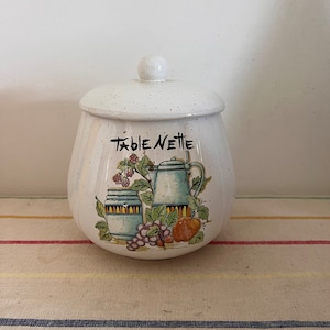 May include: White ceramic jar with a lid, featuring a speckled glaze. The jar has a hand-painted design of a teapot, two jars, grapes, and an apple. The text "Table Nettie" is written on the front of the jar.