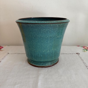 Puede incluir: Un macetero de cerámica de color verde azulado con un borde marrón. El macetero tiene un esmalte moteado y un labio ligeramente acampanado.