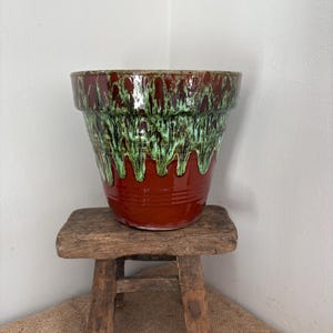 Puede incluir: Una maceta de cerámica con acabado brillante, que presenta un color base marrón rojizo intenso y un efecto decorativo de esmalte goteado en tonos verde claro y verde oscuro que cae desde el borde hacia el centro del recipiente.