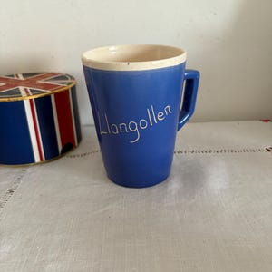 Puede incluir: Una taza de cerámica azul con un borde y un asa blancos. La taza tiene la palabra "Llangollen" escrita en blanco en el lateral.