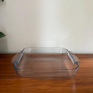 May include: A clear, square glass baking dish with a rectangular base and slightly curved sides. The dish has two handles on either side. The dish is sitting on a wooden surface.