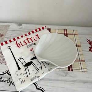 May include: White ceramic shell-shaped dish on a white and red checkered tablecloth with a napkin featuring a black and white illustration of a Parisian bistro with the text "Bistrot Paris".