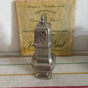 Könnte beinhalten: Ein silberfarbener, antiker Salzstreuer mit dekorativer Spitze und einer Reihe rautenförmiger Löcher. Der Streuer hat einen achteckigen Sockel und Körper. Dahinter eine Vintage-Champagner-Werbung.