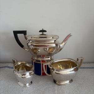 Può includere: Un servizio da tè in argento composto da una teiera, una lattiera e una zuccheriera. La teiera ha un manico nero e un coperchio con un piccolo pomello nero. La lattiera e la zuccheriera hanno manici. Il set è esposto su una superficie bianca.