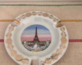 Könnte beinhalten: Weiße Porzellan-Aschenbecher mit goldfarbenem Rand und floralem Design. In der Mitte befindet sich ein farbiges Bild des Eiffelturms mit dem Text "Paris Souvenir de la Tour Eiffel".
