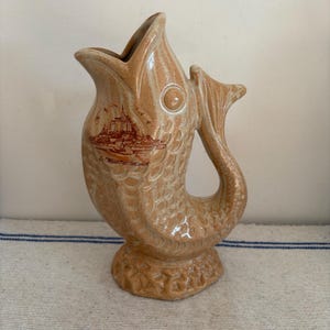 Puede incluir: Jarra de cerámica de color beige con forma de pez, con una boca ancha y una asa de cola. La jarra tiene un cuerpo texturizado y escamoso y una ilustración marrón de un edificio con texto. La base tiene un diseño decorativo y texturizado.