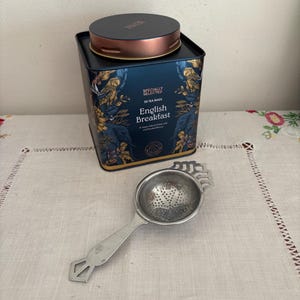 Puede incluir: Una lata de té azul y dorada con el texto "Specially Selected 50 Tea Bags English Breakfast" y un colador de té plateado.
