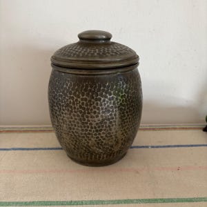 May include: A silver-coloured, hammered metal jar with a lid. The jar has a round body and a slightly flared top. The lid is also hammered and has a small knob on top.