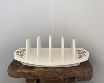 May include: A white ceramic toast rack with five vertical slots for holding slices of bread. The base features a decorative floral pattern on both the left and right sides, consisting of small yellow and orange flowers with green leaves.