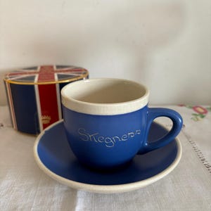 May include: A blue teacup and saucer set with the word "Stegners" in cursive script. The cup has a white rim and handle. A small, round box with a Union Jack design is in the background. The set is on a white tablecloth.