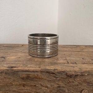 May include: A silver napkin ring with horizontal etched lines. The ring is cylindrical and sits on a weathered wooden surface. The background is a plain white wall. This decorative item is suitable for table settings.