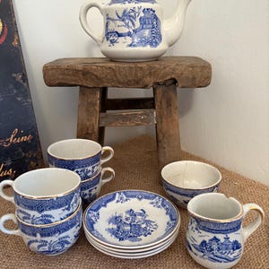 May include: A vintage tea set with blue and white landscape designs. The set includes a teapot, teacups, saucers, and a small bowl. The teapot sits on a small wooden stool. The teacups and saucers are stacked together.