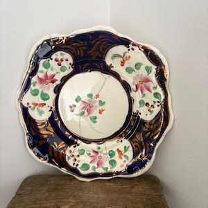 May include: An antique decorative plate with a scalloped edge. The plate has a dark blue border with gold accents and floral designs in pink, green, and white. The centre of the plate is white with a pink floral pattern.