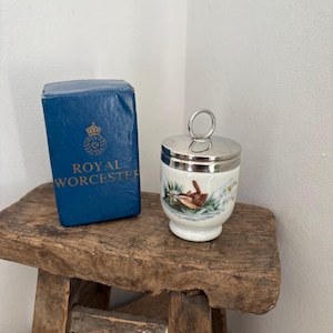 May include: A Royal Worcester egg cup with a silver lid and a painted bird scene. The egg cup is white with a detailed illustration. A blue box with the Royal Worcester logo is next to the egg cup.