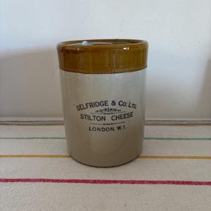 Puede incluir: Un tarro de queso cilíndrico de gres vintage con borde y base marrones. El tarro está etiquetado como "SELFRIDGE & Co. LTD. STILTON CHEESE LONDON, W.1." en texto azul. El tarro tiene un cuerpo de color crema y probablemente sea de principios del siglo XX.