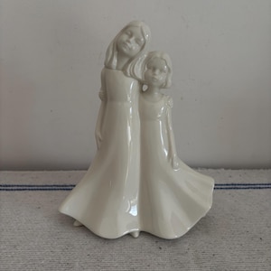 May include: A cream-colored ceramic figurine depicting two young girls embracing. The girls are wearing long dresses with flowing skirts. The figurine has a glossy finish and is set against a neutral background.