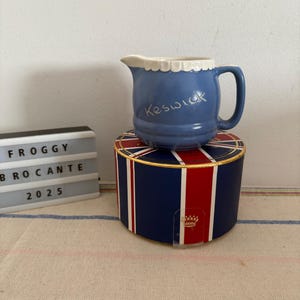 Puede incluir: Una jarra de crema de cerámica azul con un borde blanco festoneado y la palabra "Keswick" escrita en blanco en el lateral. La jarra está sobre una caja roja, blanca y azul con una corona dorada.