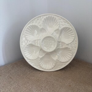 May include: A round, off-white ceramic oyster plate featuring six molded shell-shaped indentations arranged in a circle around a central circular well. The surface has a textured, pebbled finish and a rope-like border detail around the center.