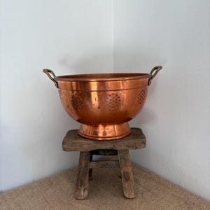 Op de afbeelding: Een grote, ronde koperen vergiet met een gepolijste metalen afwerking, twee gebogen koperen handgrepen aan de zijkanten en een brede voet. De kom is geperforeerd met kleine, gelijkmatig verdeelde ronde gaatjes in patronen.