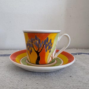 Könnte beinhalten: Ein farbenfrohes Keramik-Tassen- und Untertassen-Set. Die Tasse zeigt ein Baumdesign mit orangefarbenen, gelben und blauen Farbtönen. Die Untertasse hat ein passendes Regenbogenmuster. Das Set steht vor einem weißen Hintergrund.