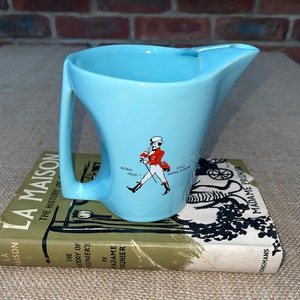 May include: A light blue ceramic jug with a handle and a spout. The jug features a graphic of a man in a red jacket and white trousers walking with a cane. The text "Born 1820" and "Still Going Strong" is printed on the jug.