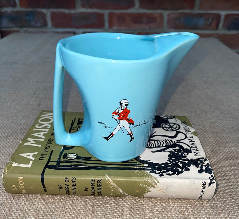 May include: A light blue ceramic jug with a handle and a spout. The jug features a graphic of a man in a red jacket and white trousers walking with a cane. The text "Born 1820" and "Still Going Strong" is printed on the jug.