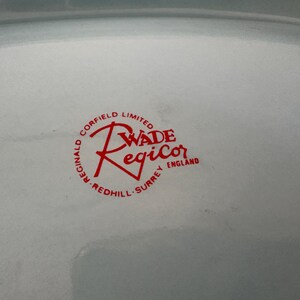 May include: Close-up of a white ceramic plate with a red logo. The logo reads "Wade Regicor England" in a circular design with "Reginald Corfield Limited" and "Redhill Surrey" around the edge.