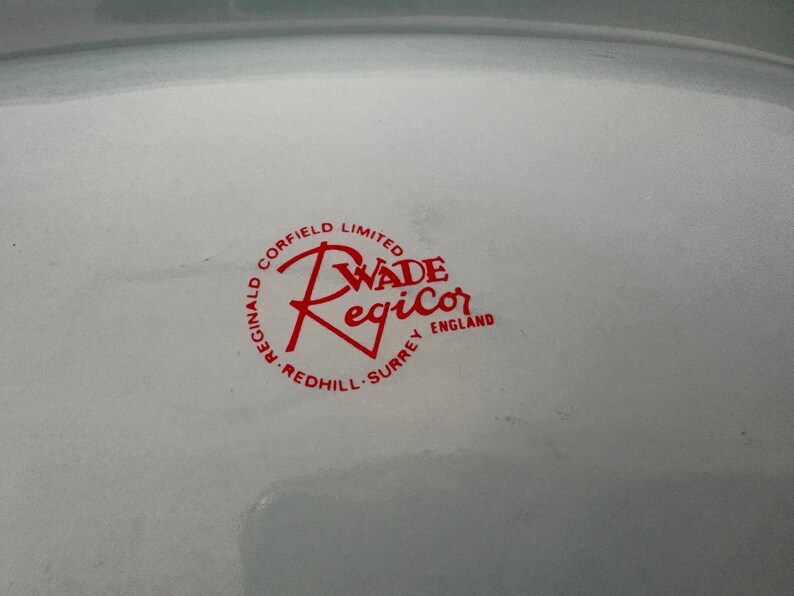 May include: Close-up of a white ceramic plate with a red logo. The logo reads "Wade Regicor England" in a circular design with "Reginald Corfield Limited" and "Redhill Surrey" around the edge.